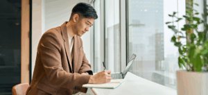 Man working with his laptop and notebook in front of large windows
