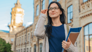 Woman in a blue shirt and gray cardigan adjusts her glasses in front of campus building.