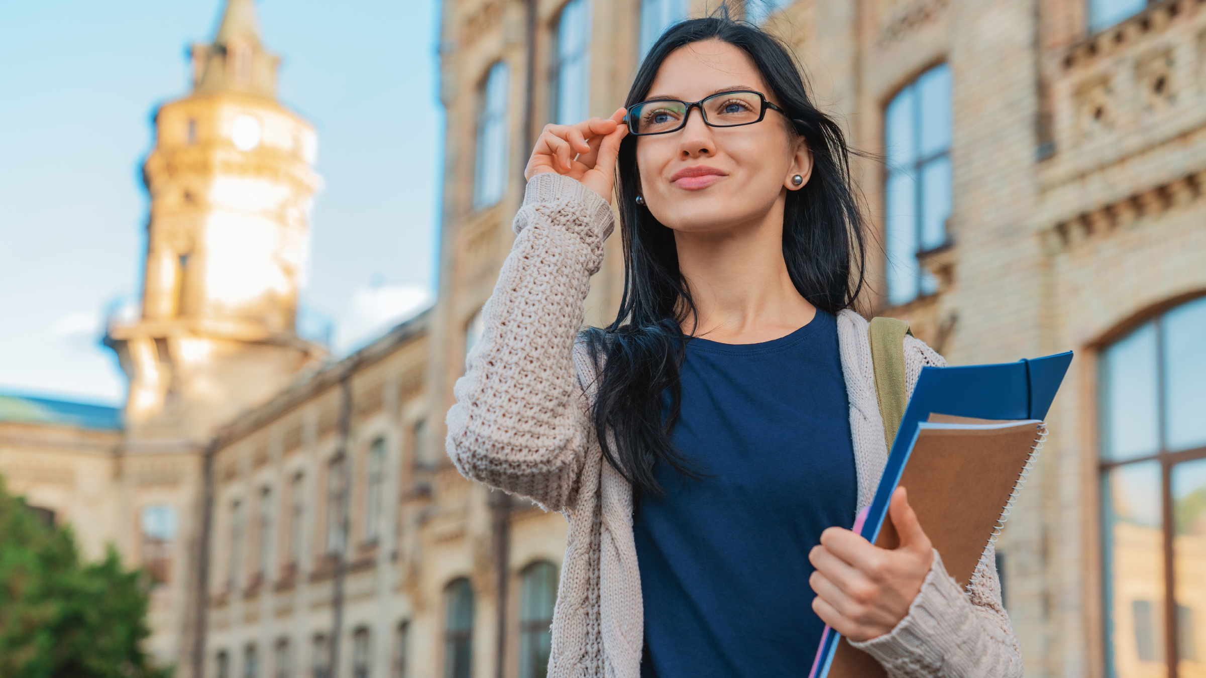 Woman in a blue shirt and gray cardigan adjusts her glasses in front of campus building.