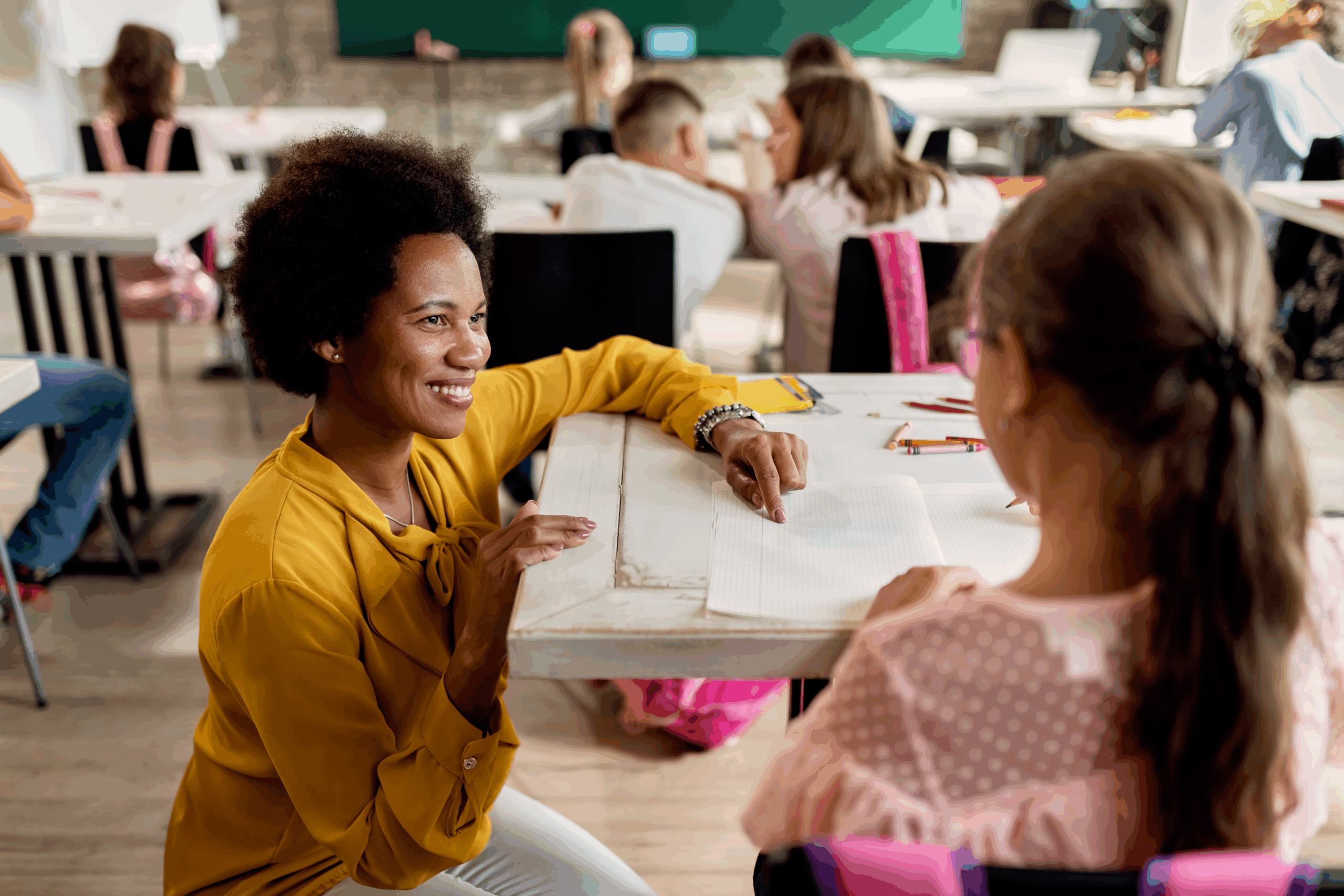 Smiling teacher in a yellow blouse kneels beside a young student in a wheelchair, helping her with a worksheet in a classroom with other children working at their desks.