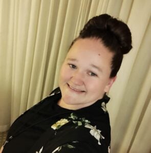 Smiling woman with brunette top knot wears black floral blouse near curtain